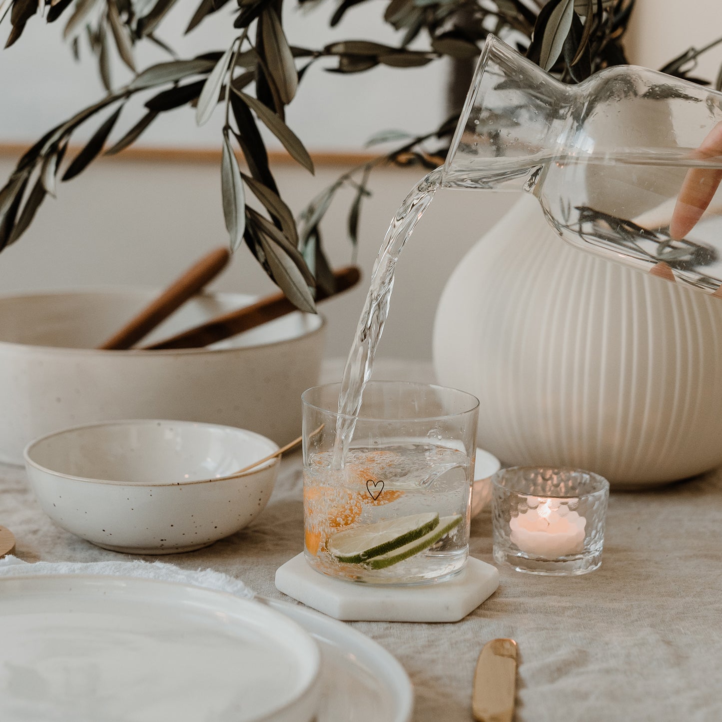 Wasser wird in Glas mit Herzmotiv und Zitrusfrüchten gegossen, auf gedecktem Tisch mit Kerze, Vase und Keramik.