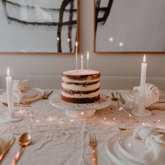 Naked Cake mit Kerzen auf weißer Tortenplatte, umgeben von Lichterkette und gedecktem Tisch.
