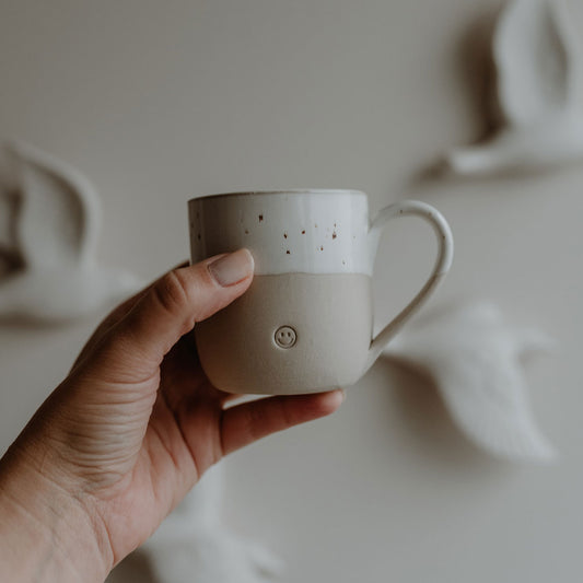 Hand hält eine beige-weiße Keramiktasse mit gesprenkelter Glasur und Henkel. Auf der Vorderseite ist ein kleines, rundes Smiley-Symbol eingeprägt. Im Hintergrund sind unscharf Wanddekorationen in Form fliegender Vögel zu sehen – das Bild vermittelt Leichtigkeit und Freude, perfekt als detailverliebte Geschenkidee.