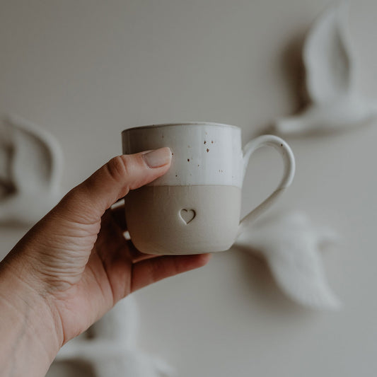 ine linke Hand hält eine handgefertigte Keramiktasse in natürlichen Beige- und Weißtönen vor einer Wand mit weißen, fliegenden Vogelskulpturen. Die obere Hälfte der Tasse ist glasiert und gesprenkelt, die untere ist matt und trägt eine kleine Herzprägung.