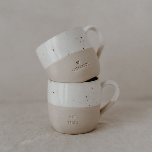 Eine beige-weiße Keramiktasse liegt auf der Seite auf einem Leinentuch. Die obere Hälfte ist glasiert und weiß mit kleinen braunen Sprenkeln, die untere Hälfte matt beige. Ein kleines, eingraviertes Herz ist an der Seite zu sehen. Auf dem Tassenboden steht in einem Kreis: ‚Zuhause bei mir‘.