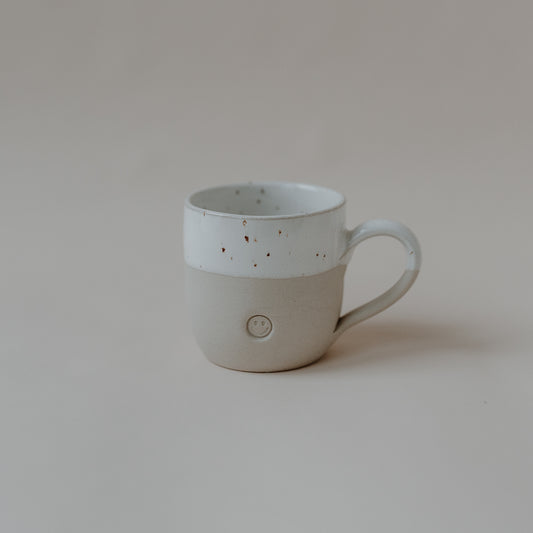 Handgefertigte Keramiktasse mit dezentem Farbverlauf von Naturbeige im unteren Bereich zu gesprenkeltem Weiß im oberen Bereich. Auf der Vorderseite ist ein kleines, rundes Relief mit einem lächelnden Smiley eingearbeitet. Die Tasse steht auf hellem Untergrund in minimalistischer Umgebung.