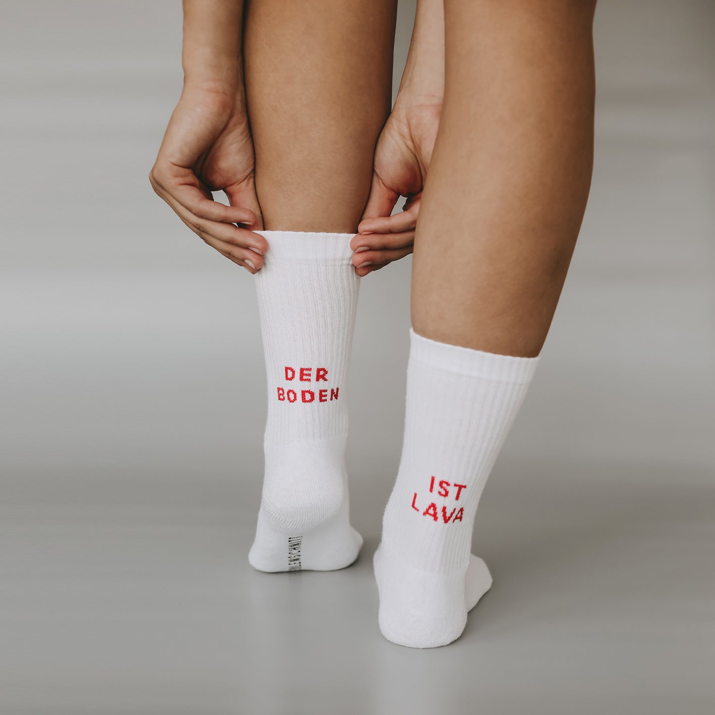 Eine Person zieht sich ihre weißen Socken hoch, auf denen in leuchtend roter Schrift steht: „DER BODEN IST LAVA“. Die Szene ist bewusst schlicht gehalten – ein neutraler Hintergrund lenkt den Fokus ganz auf die Socken und die Botschaft.