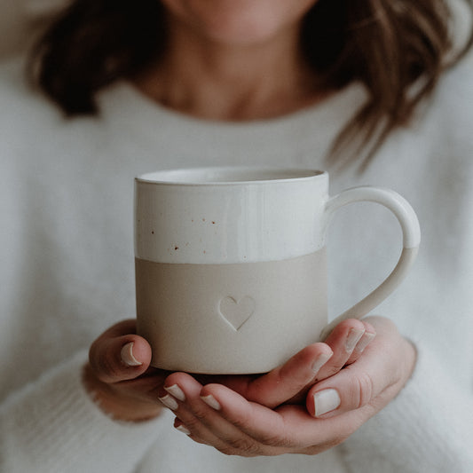 Zwei Hände halten eine beige-weiße Keramiktasse mit gesprenkelter Glasur und einem eingeprägten Herzsymbol auf der Vorderseite. Die Person trägt einen flauschigen, weißen Pullover – das Bild strahlt Wärme, Achtsamkeit und liebevolle Verbundenheit aus.