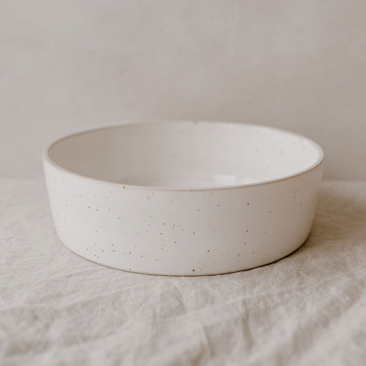 Große, flache Schale aus heller Keramik mit feiner Sprenkelung, auf einem Leinentuch arrangiert. Die minimalistische Formensprache und natürliche Oberfläche stehen im Fokus.