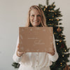 Lächelnde Frau hält die limitierte Winter-Überraschungsbox 2026 von Eulenschnitt mit liebevollen Wintermotiven vor einem beleuchteten Weihnachtsbaum.