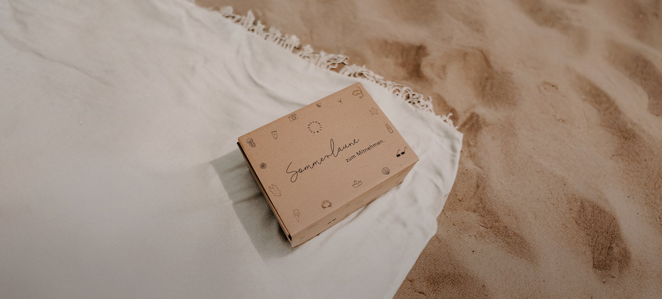 Sommerbox „Sommerlaune zum Mitnehmen“. Erhältlich am 20. August um 18 Uhr für 45 €. Abgebildet ist die Sommerüberraschungsbox auf einer hellen Decke am Sandstrand. Hinweis: zzgl. Versandkosten, solange der Vorrat reicht.