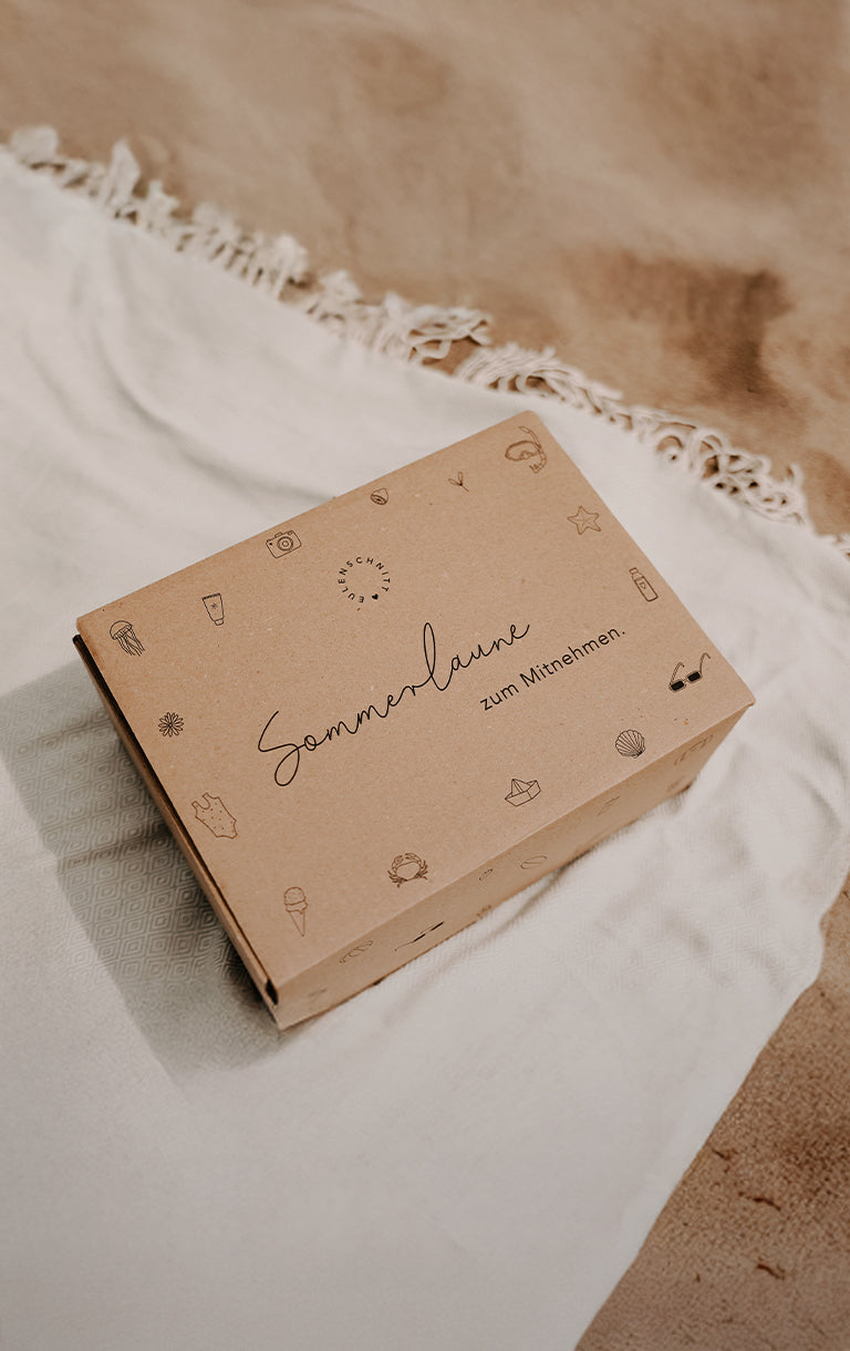 Sommerbox „Sommerlaune zum Mitnehmen“. Erhältlich am 20. August um 18 Uhr für 45 €. Abgebildet ist die Sommerüberraschungsbox auf einer hellen Decke am Sandstrand. Hinweis: zzgl. Versandkosten, solange der Vorrat reicht.