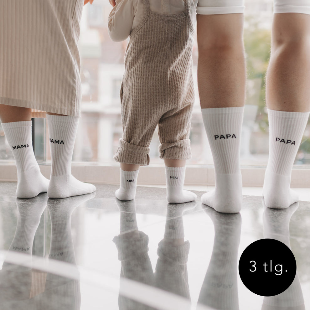 Familiensocken-Bundle in Weiß mit Aufdruck Mama, Papa und Mini – drei Paar Socken an Füßen von Eltern und Kind auf glänzendem Boden.