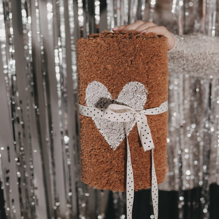 Braune Fußmatte mit weißem Herz und dekorativer Herzchen-Schleife, von einer Person vor einem glitzernden silbernen Hintergrund gehalten.