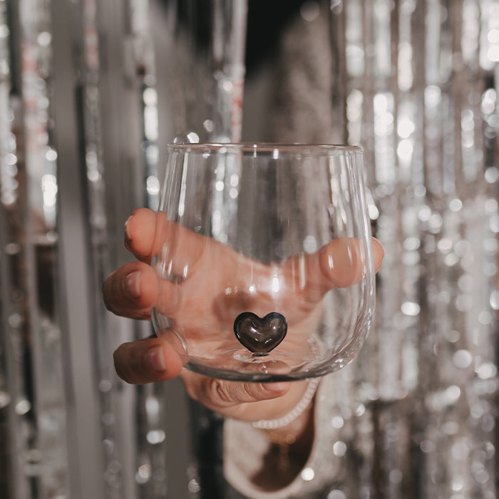 Transparentes Trinkglas mit kleiner schwarzer Herzfigur im Inneren, von einer Hand vor einem glitzernden silbernen Partyhintergrund gehalten.