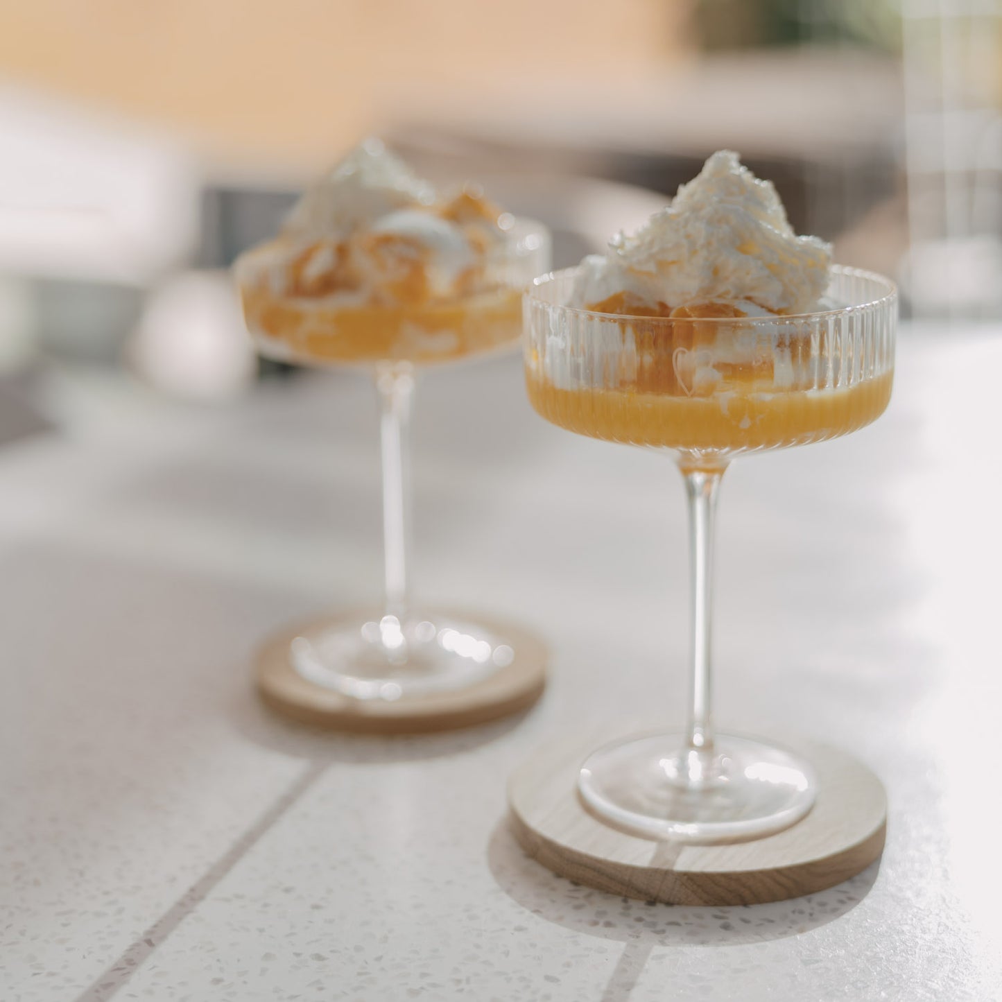 Zwei Champagnergläser mit Dessert und Sahne auf Holzuntersetzern im Sonnenlicht auf hellem Tisch.