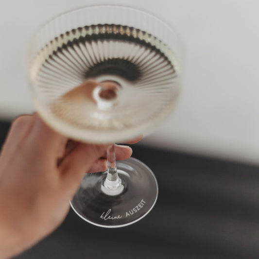 Hand hält geriffeltes Champagnerglas mit Gravur „kleine Auszeit“ – eleganter Moment zum Anstoßen.