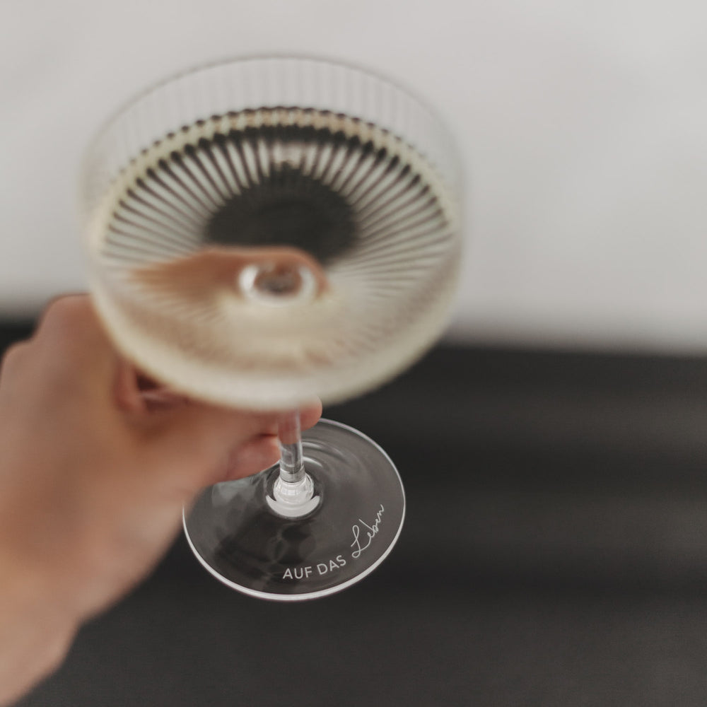 Hand hält Champagnerglas mit Gravur „Auf das Leben“ in Nahaufnahme, Fokus auf Glasboden.