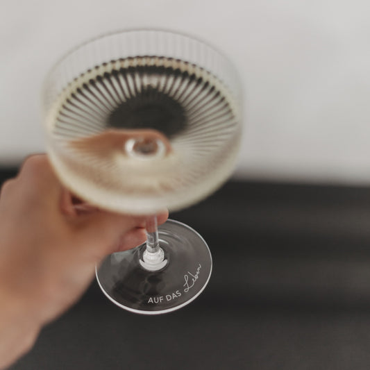 Hand hält Champagnerglas mit Gravur „Auf das Leben“ in Nahaufnahme, Fokus auf Glasboden.