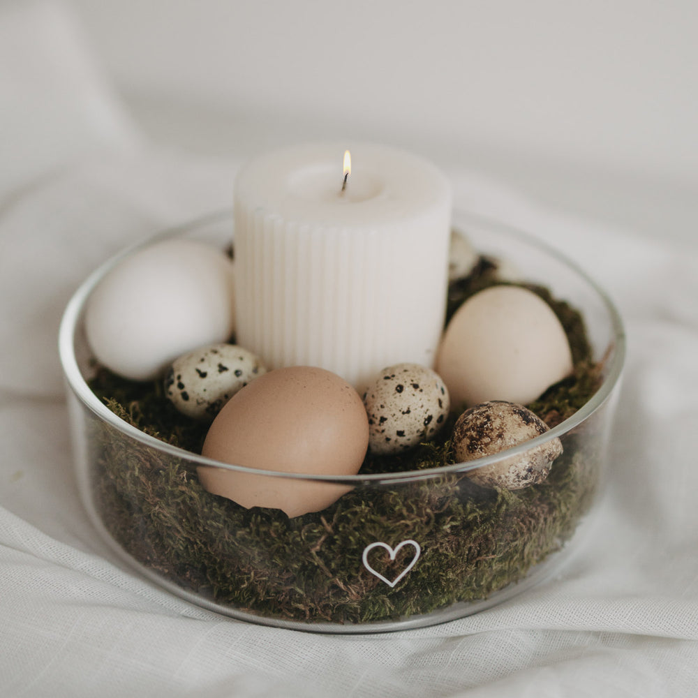 Glas-Schale mit Herzmotiv, dekoriert mit Moos, Eiern und weißer Stumpenkerze als natürliche Osterdekoration.