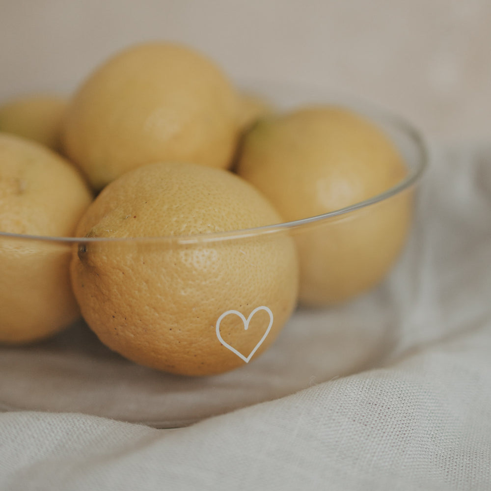 Glas-Schale mit weißem Herzmotiv, gefüllt mit Zitronen auf einem Stofftuch als natürliche Küchendeko.