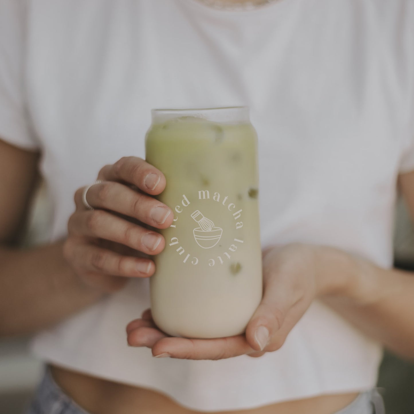 Zwei Hände halten ein Glas mit eisgekühltem Matcha Latte. Das minimalistische Glas mit Beschriftung "Iced Matcha latte club" steht vor einer lässig in Jeans und weißes Shirt stehenden Frau.