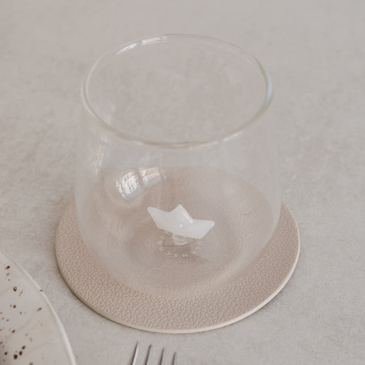 Trinkglas mit Papierboot-Motiv innen, auf Untersetzer neben Teller und Besteck, minimalistisches Setting