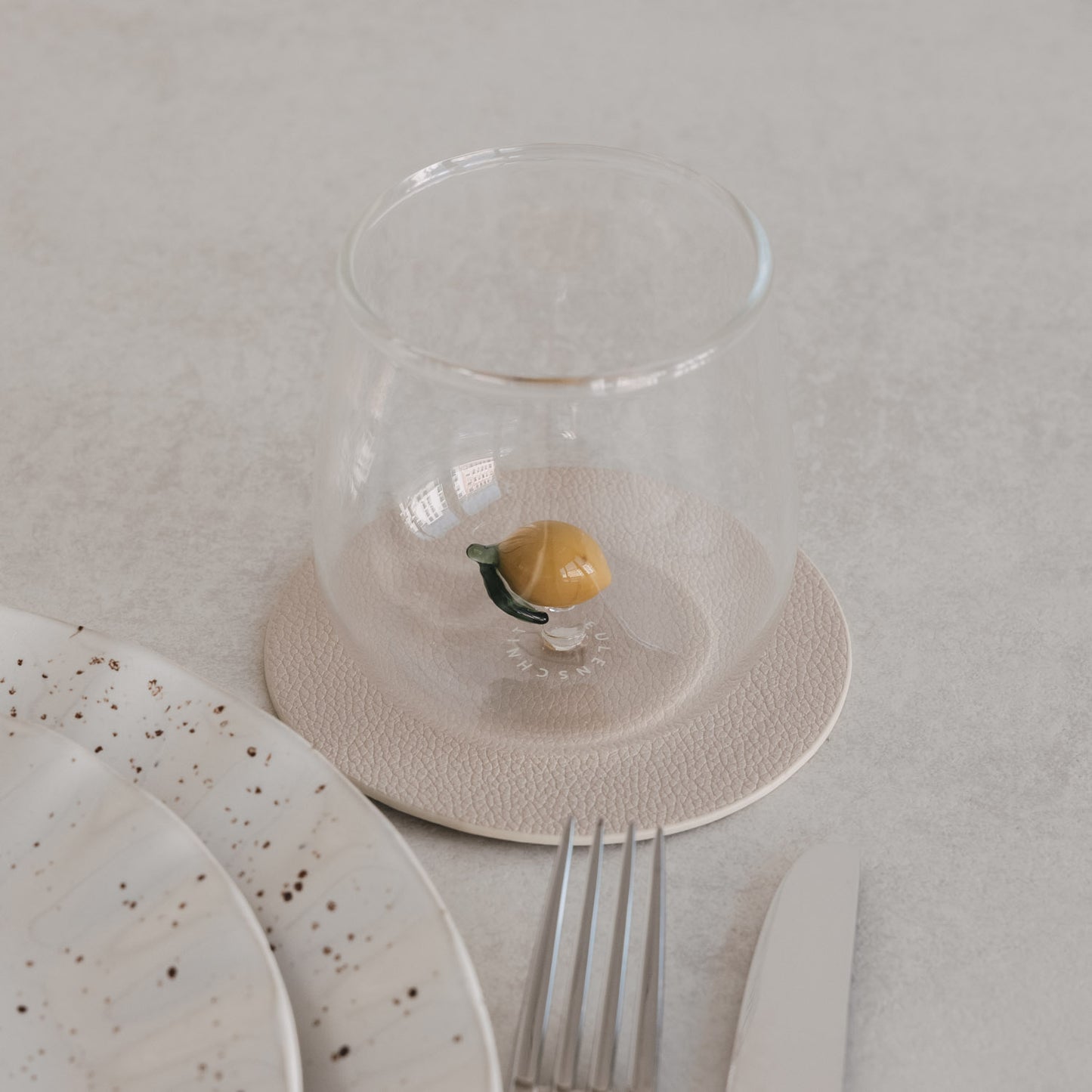 Glas mit Zitronenmotiv innen, auf Untersetzer neben Teller und Besteck, minimalistisches Tischsetting