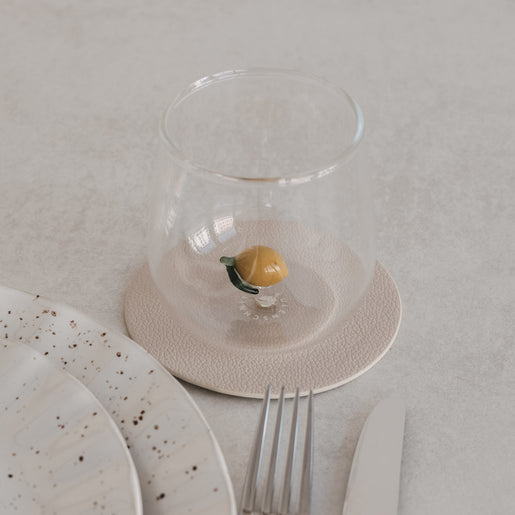 Glas mit Zitronenmotiv innen, auf Untersetzer neben Teller und Besteck, minimalistisches Tischsetting