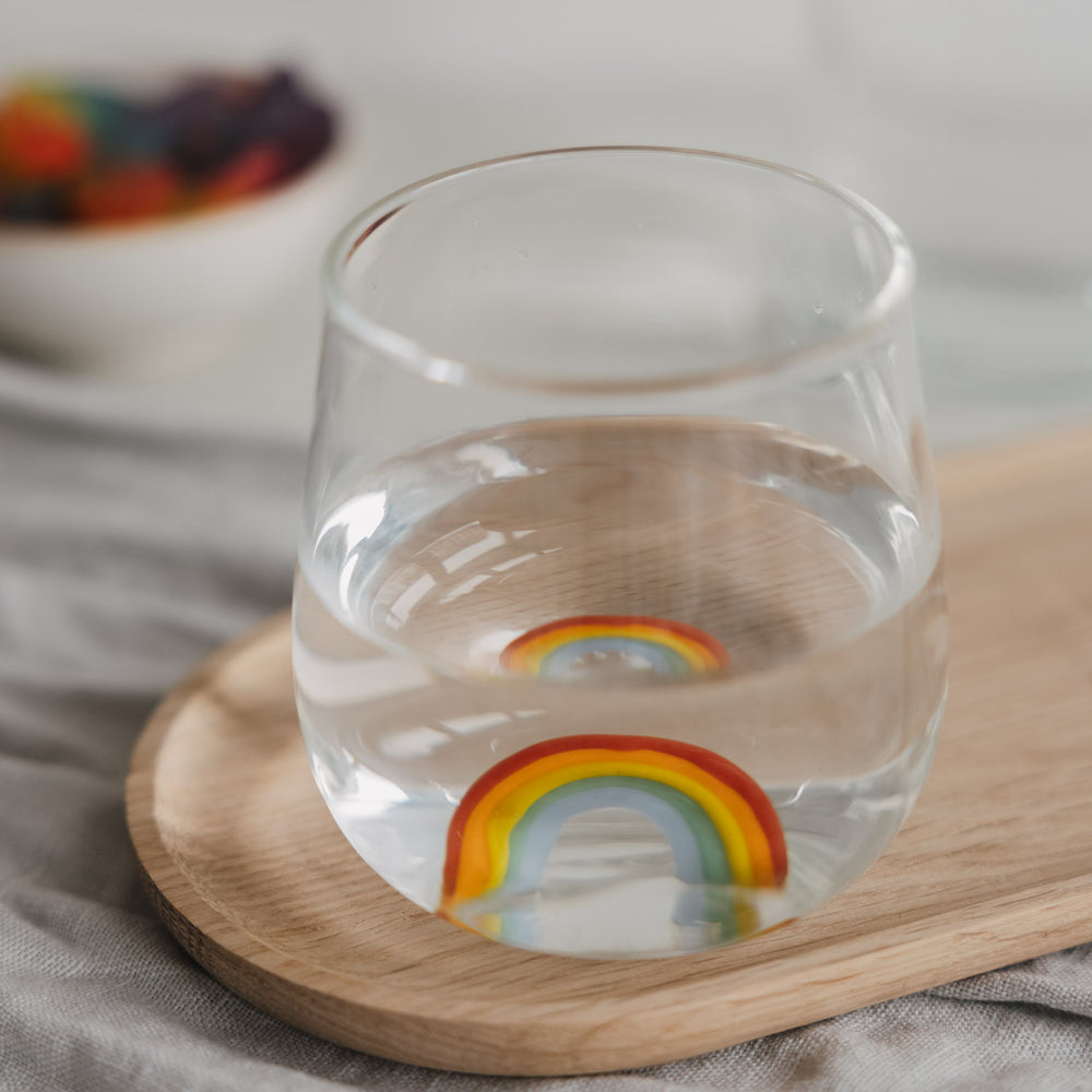 Trinkglas mit Regenbogen Motiv gefüllt mit Wasser, dekorativ auf Holzbrett arrangiert