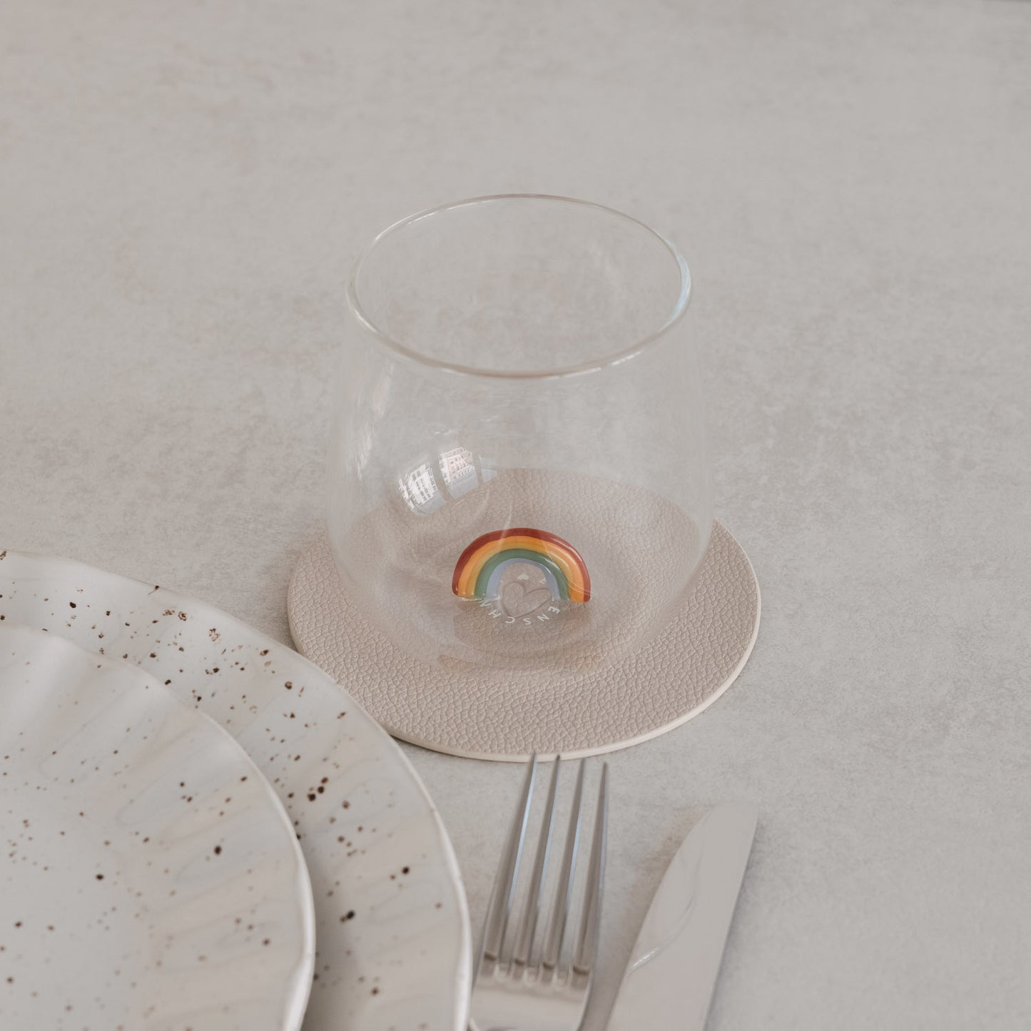 Trinkglas mit Regenbogen-Motiv innen, auf Untersetzer neben Teller und Besteck, minimalistisches Setting