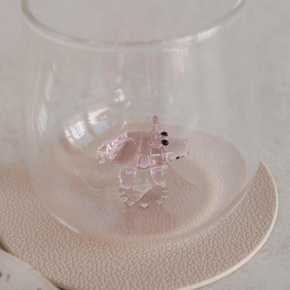 Trinkglas mit rosa Schwein-Figur innen, auf Untersetzer, modernes minimalistisches Tischsetting