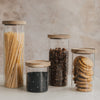 Vier Vorratsgläser mit kleinen weißen Herzen und Holzdeckeln, gefüllt mit Spaghetti, Hülsenfrüchten, Müsli und Keksen.