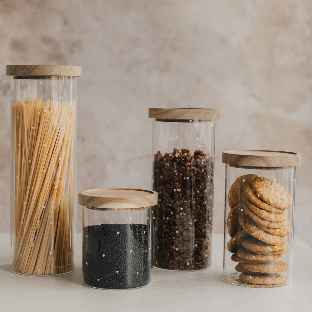 Vier Vorratsgläser mit kleinen weißen Herzen und Holzdeckeln, gefüllt mit Spaghetti, Hülsenfrüchten, Müsli und Keksen.