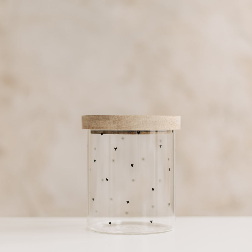 Leeres Vorratsglas mit kleinen schwarzen Herzen und aufgesetztem Holzdeckel vor hellem Hintergrund.