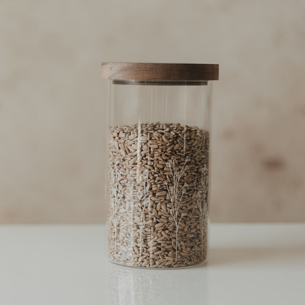 Hoher Glasbehälter mit weißem Wildblumen-Motiv und Holzdeckel, gefüllt mit Sonnenblumenkernen
