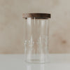 Leerer hoher Glas-Vorratsbehälter mit filigranem weißem Wildblumen-Motiv und dunklem Holzdeckel