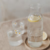 Glas Karaffe mit Zitronenwasser und zwei Gläsern mit Blüten auf Holztablett