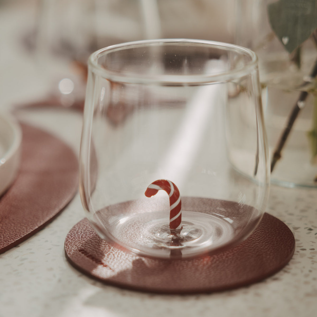 Transparentes Figurenglas mit rot-weißer Zuckerstange im Glasboden auf einem braunen Untersetzer.