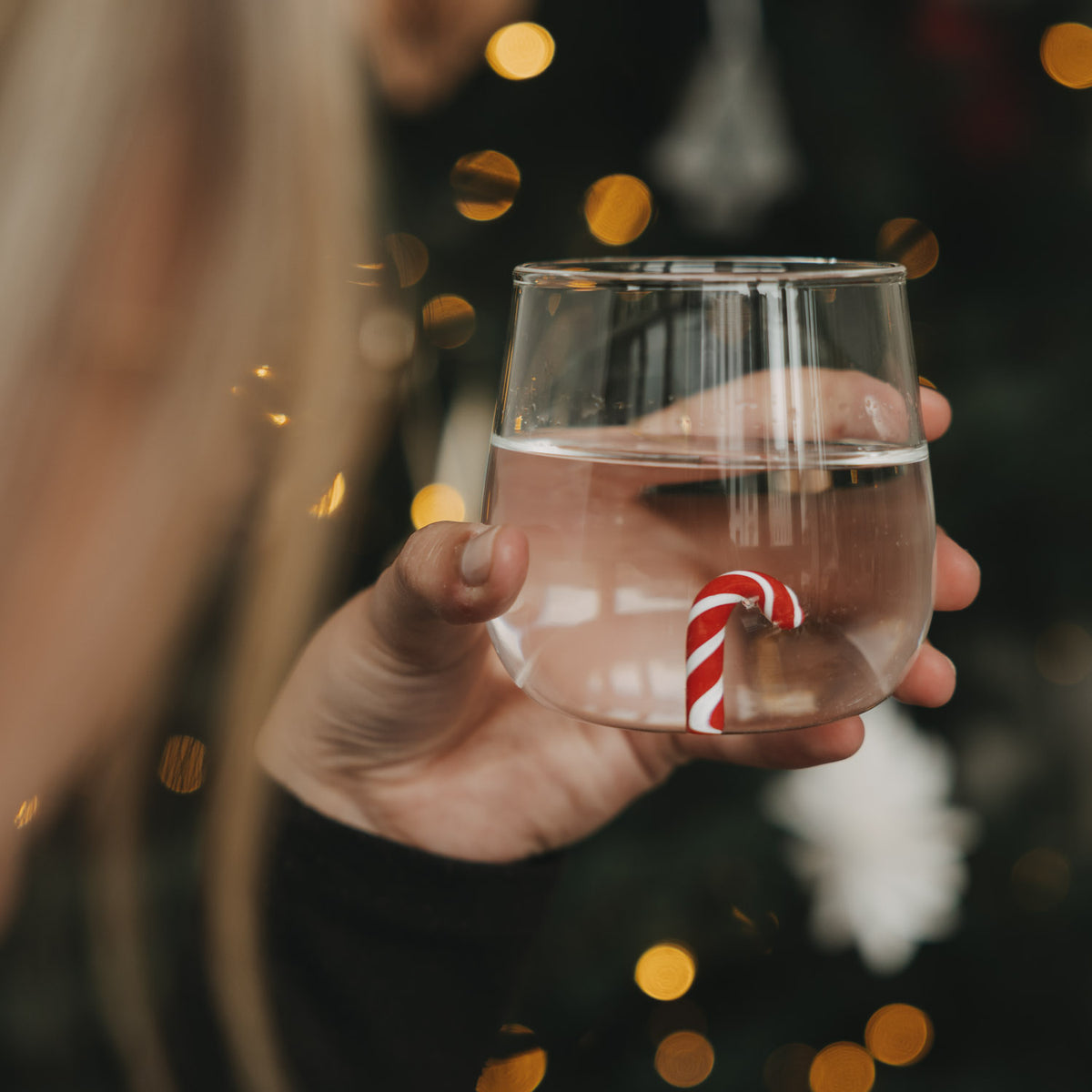 Hand hält ein Figurenglas mit Zuckerstange vor einem weihnachtlich beleuchteten Hintergrund.