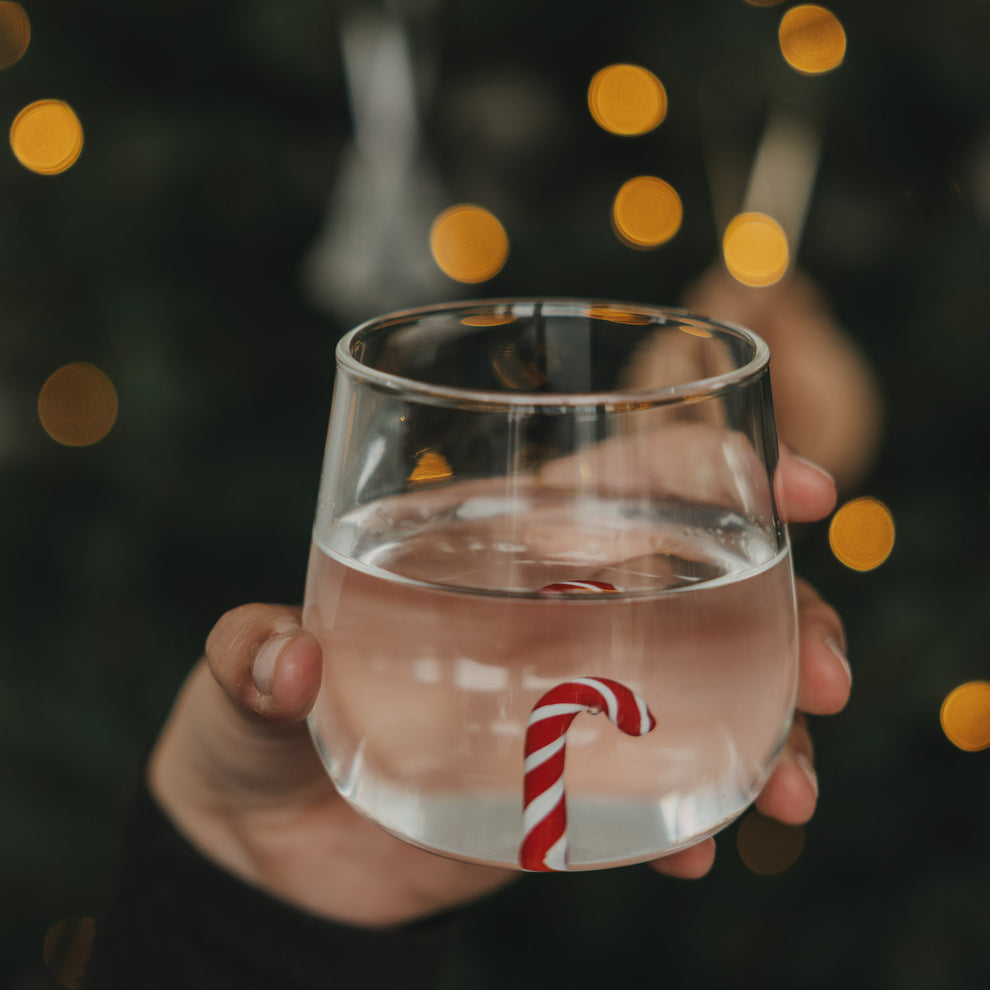 Transparentes Trinkglas mit rot-weißer Zuckerstange im Glasboden, gefüllt mit Wasser vor warmen Weihnachtslichtern.