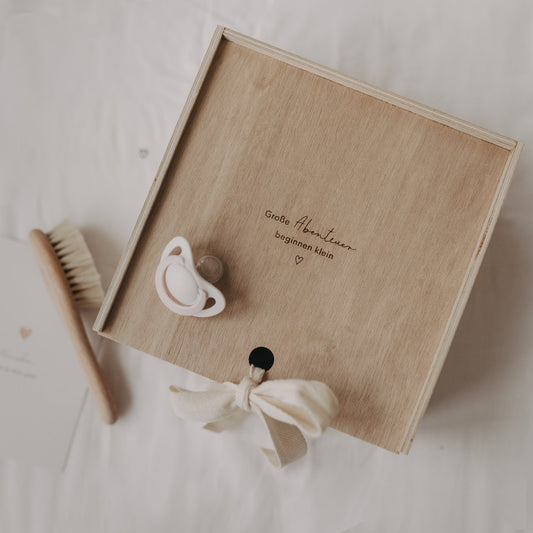 Große Erinnerungsbox aus Holz mit Gravur „Große Abenteuer beginnen klein“, Schnuller und Babybürste auf hellem Untergrund.
