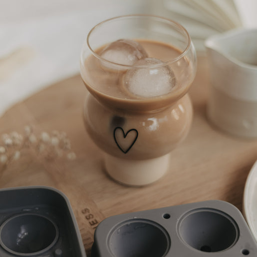 Eulenschnitt Bubble Glas mit schwarzem Herz-Print, gefüllt mit Iced Coffee und runden Eiswürfeln. Davor eine Silikoneiswürfelform auf einem runden Holzbrett.