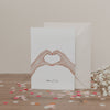 Klappkarte mit zwei Händen in Herzform und dem Schriftzug „Alles Liebe“, vor Umschlag und Blumen, auf hellem Untergrund.
