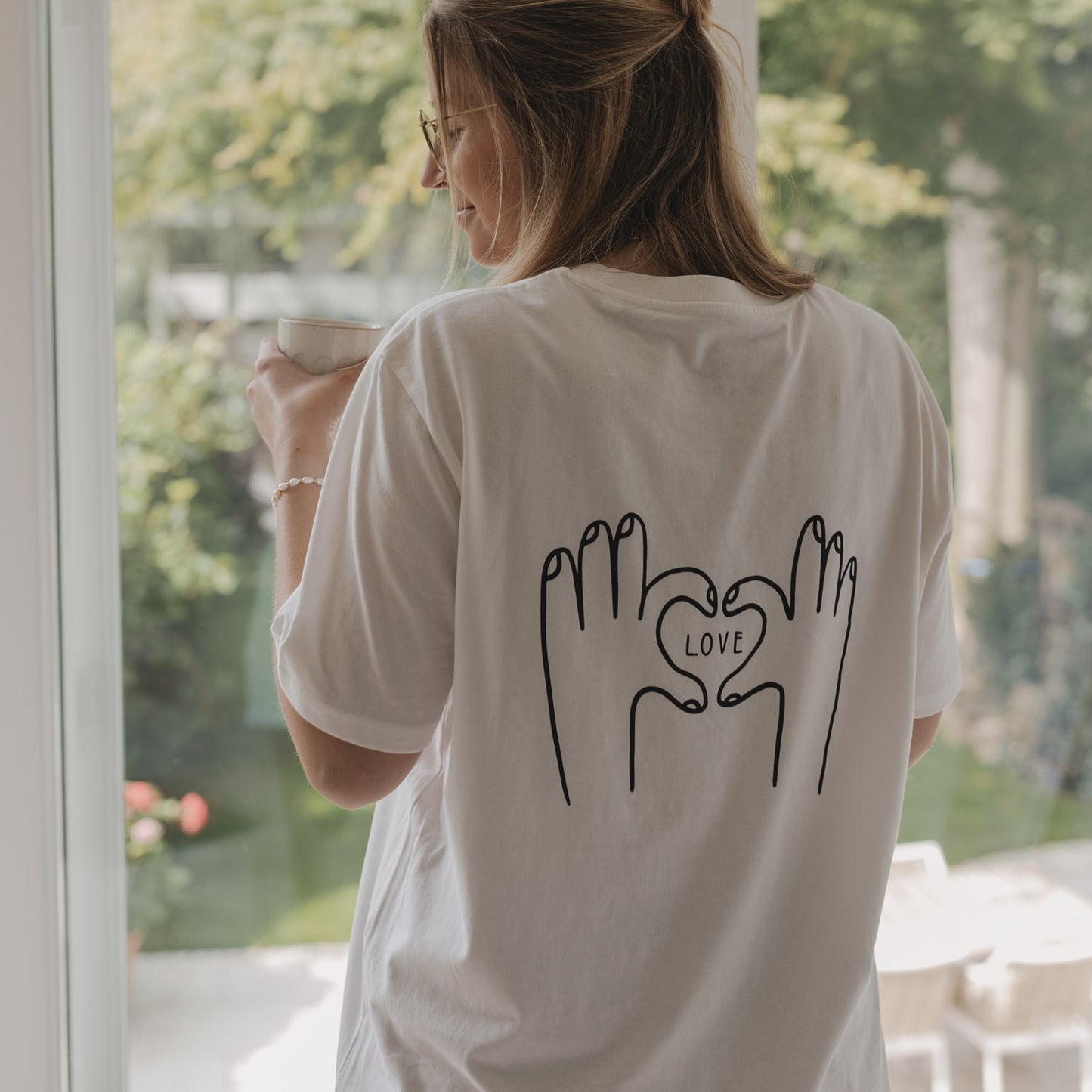 Frau mit Brille und LOVE-T-Shirt steht mit Kaffeetasse am Fenster