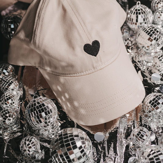 Beige Cap mit kleinem schwarzem Herz auf der Vorderseite, dekoriert zwischen silbernen Discokugeln und glänzender Partyfolie.
