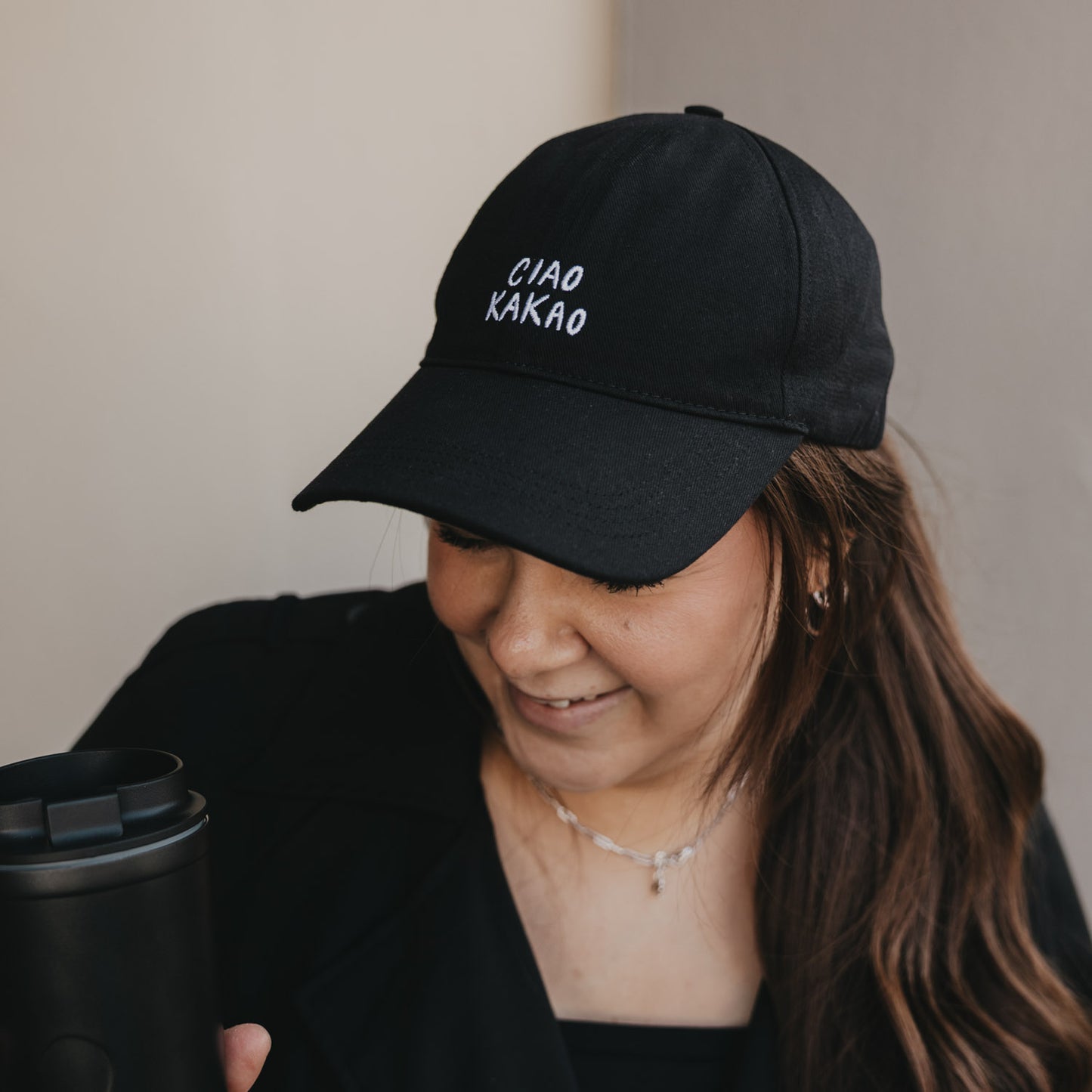 Frau trägt schwarze Cap mit weißer Stickerei „Ciao Kakao“ und hält Coffee-to-go Becher
