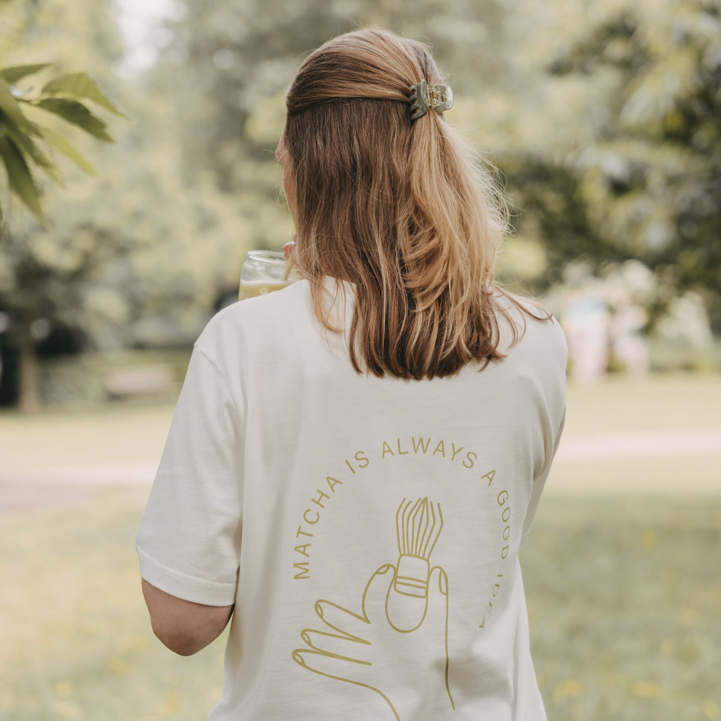 Frau trägt weißes Matcha T-Shirt und hält Getränk im Park, Rückenansicht