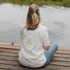 Frau mit weißem Matcha T-Shirt am See, Rückenansicht mit Getränk