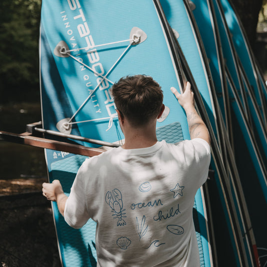 Mann mit weißem „Ocean Child“ T-Shirt beim Tragen von Paddleboards, aktiver Lifestyle