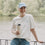 Frau mit weißem Coffee Club T-Shirt und Becher am Wasser, entspannter Lifestyle