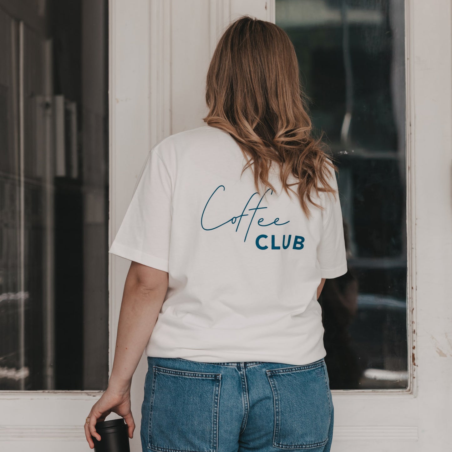 Rückansicht: weißes T-Shirt mit „Coffee Club“ Schriftzug, Frau mit Becher vor Haustür