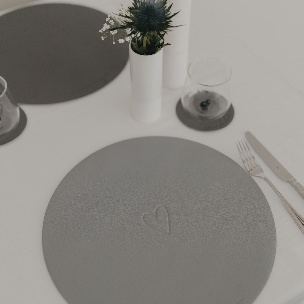 Hellgraues rundes Platzset mit eingeprägtem Herzmotiv, dekoriert mit Vase und Besteck auf weißem Tisch.