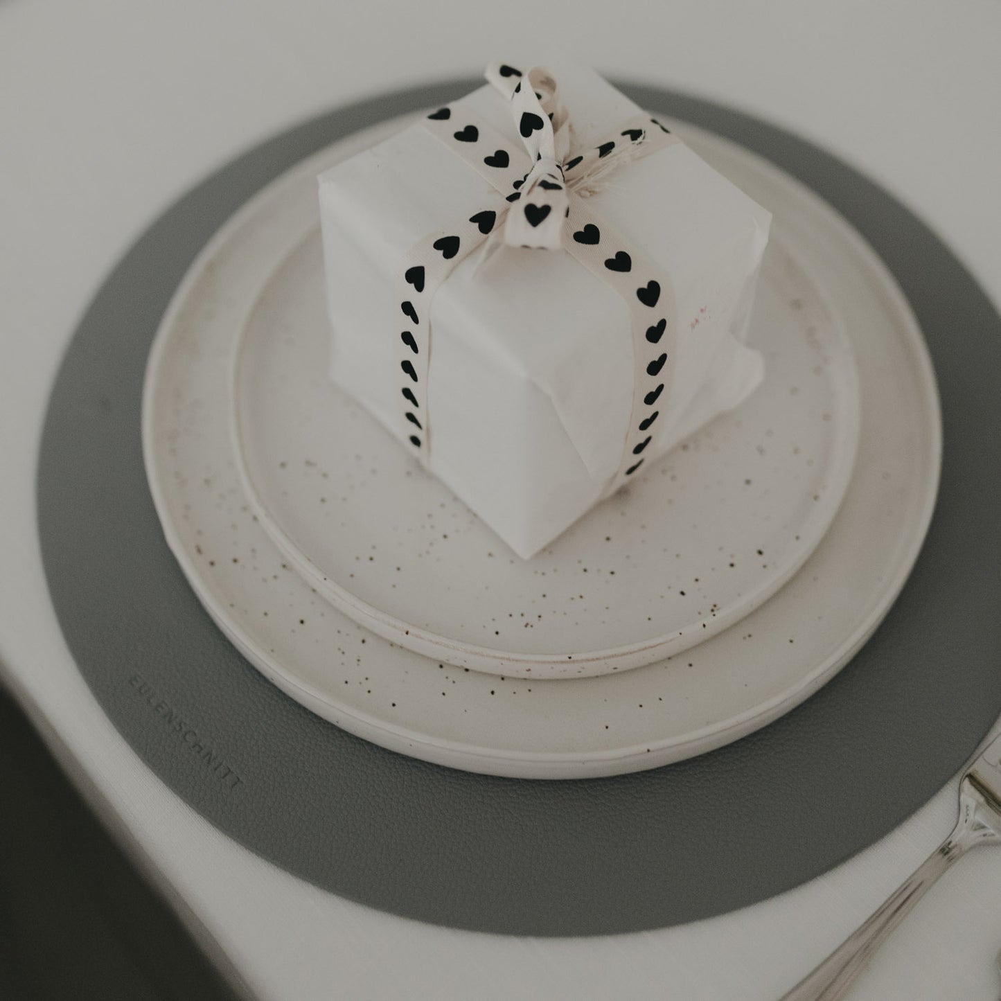 Nahaufnahme eines grauen Platzsets mit weißem Geschirr und herzverziertem Geschenkband.